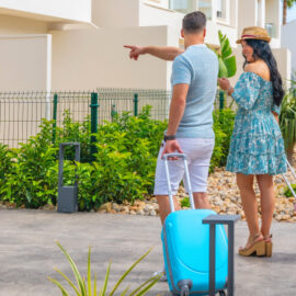 Couple with rolling suitcases walking toward a modern vacation rental property on a sunny day, illustrating short-term rental real estate.